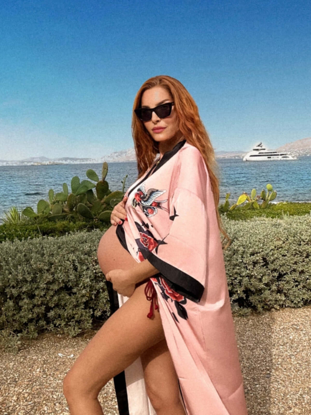 Woman wearing a Rose Butterfly Kimono Robe by the water, showcasing luxury and comfort during pregnancy.
