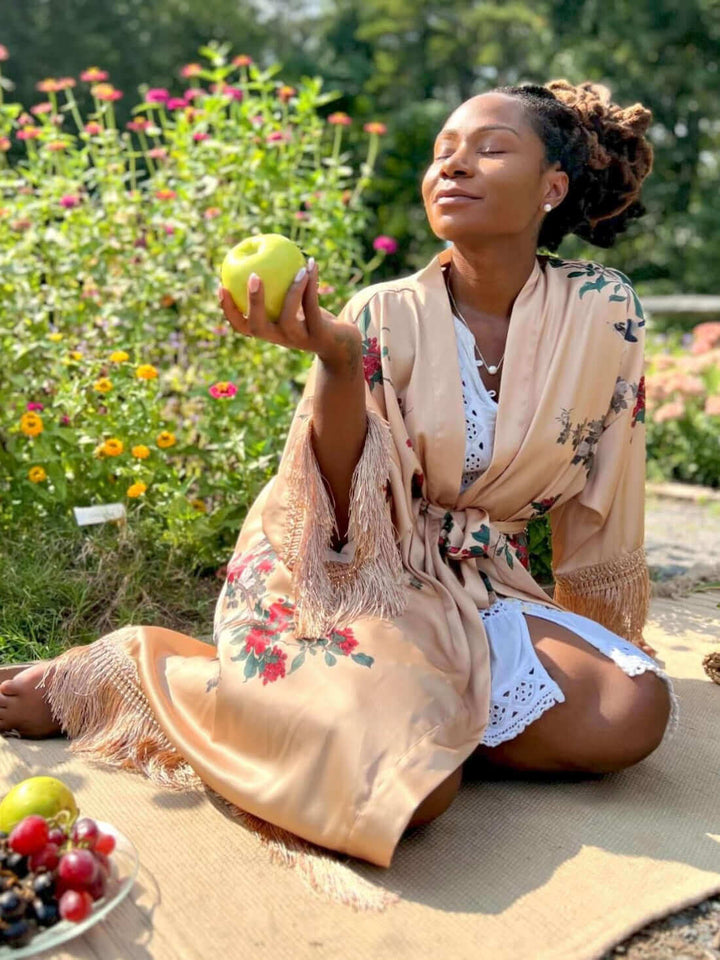 Woman in a Tassels Champagne Kimono Robe holding an apple surrounded by colorful flowers.