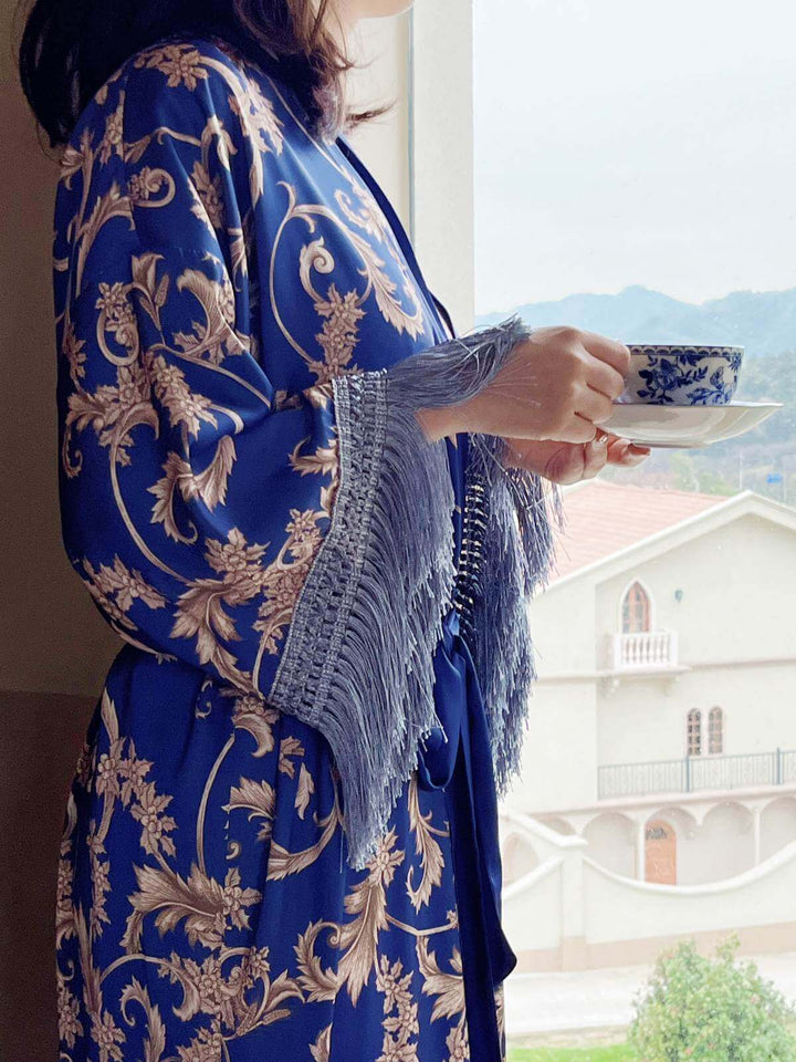 Woman wearing a Tassels Blue Kimono Robe, holding a teacup by the open window, showcasing luxurious fabric and design.