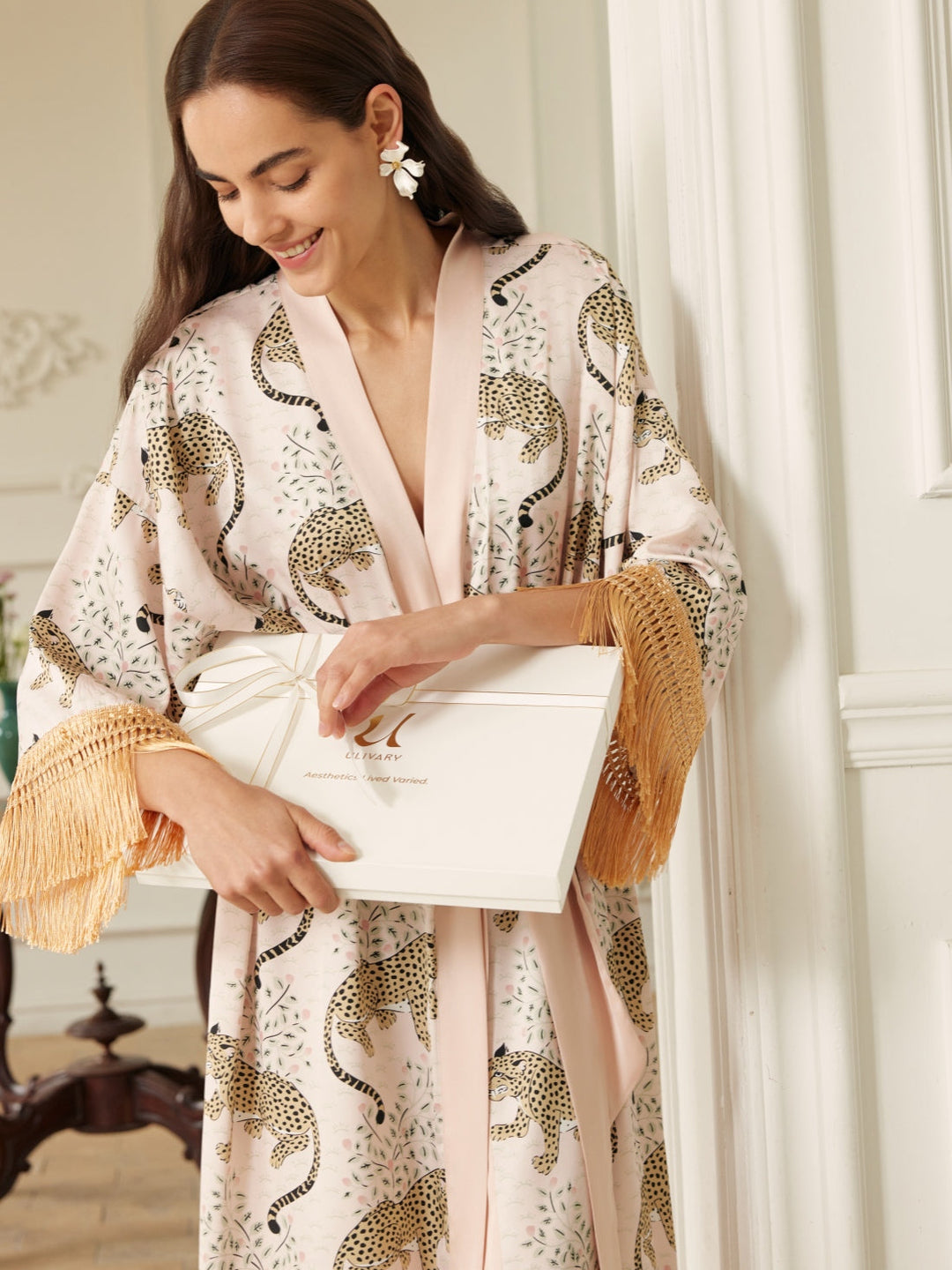 Woman wearing a Pink Leopard Tassel Kimono Robe with tassel sleeves, holding a gift box in a stylish setting.