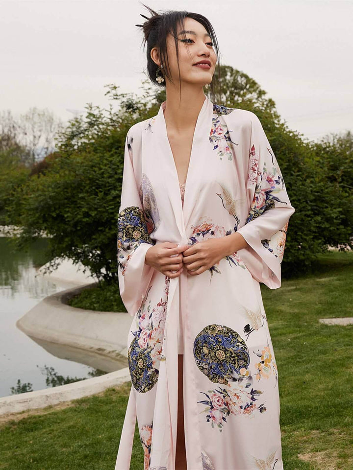 Woman wearing a Garden Crane Blush Kimono Robe showcasing hand-painted floral designs near a serene garden pond.