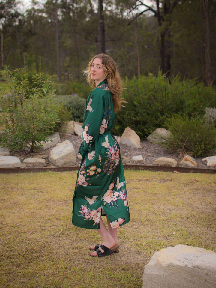 A woman wearing a Garden Crane Green Kimono Robe in a garden, showcasing luxury and elegant floral designs.