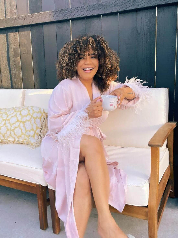 Woman in Pink Feather Bridal Robe sitting on a sofa, enjoying a cup of tea in a luxurious setting.